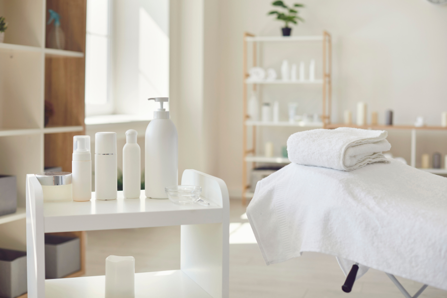 White sheet draped over massage table with white towel and white bottles on shelf